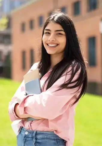 A smiling teenage girl with long dark hair holding notebooks outside on a sunny day